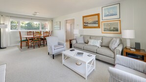Ein Wohnzimmer mit Essbereich, einer Couch, einem Couchtisch und einer Lampe. An den Wänden hängen gerahmte Bilder.