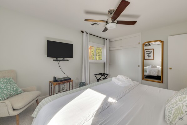 Habitación de hotel con cama, ventilador de techo, televisión, ventana con cortinas, espejo y una mesita de noche con lámpara.