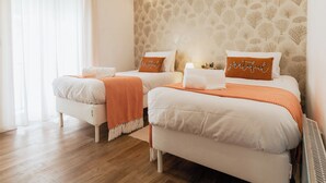 Ein Hotelzimmer mit zwei Betten, jeweils mit weißer Bettwäsche und orangefarbenen Ziertelementen, Holzboden und Wandtapete mit Muster.