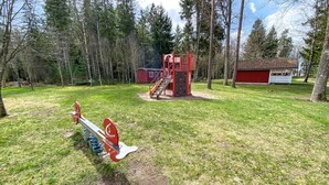 Ein Spielplatz mit Wippe und Klettergerüst, umgeben von Bäumen und einem roten Gebäude.