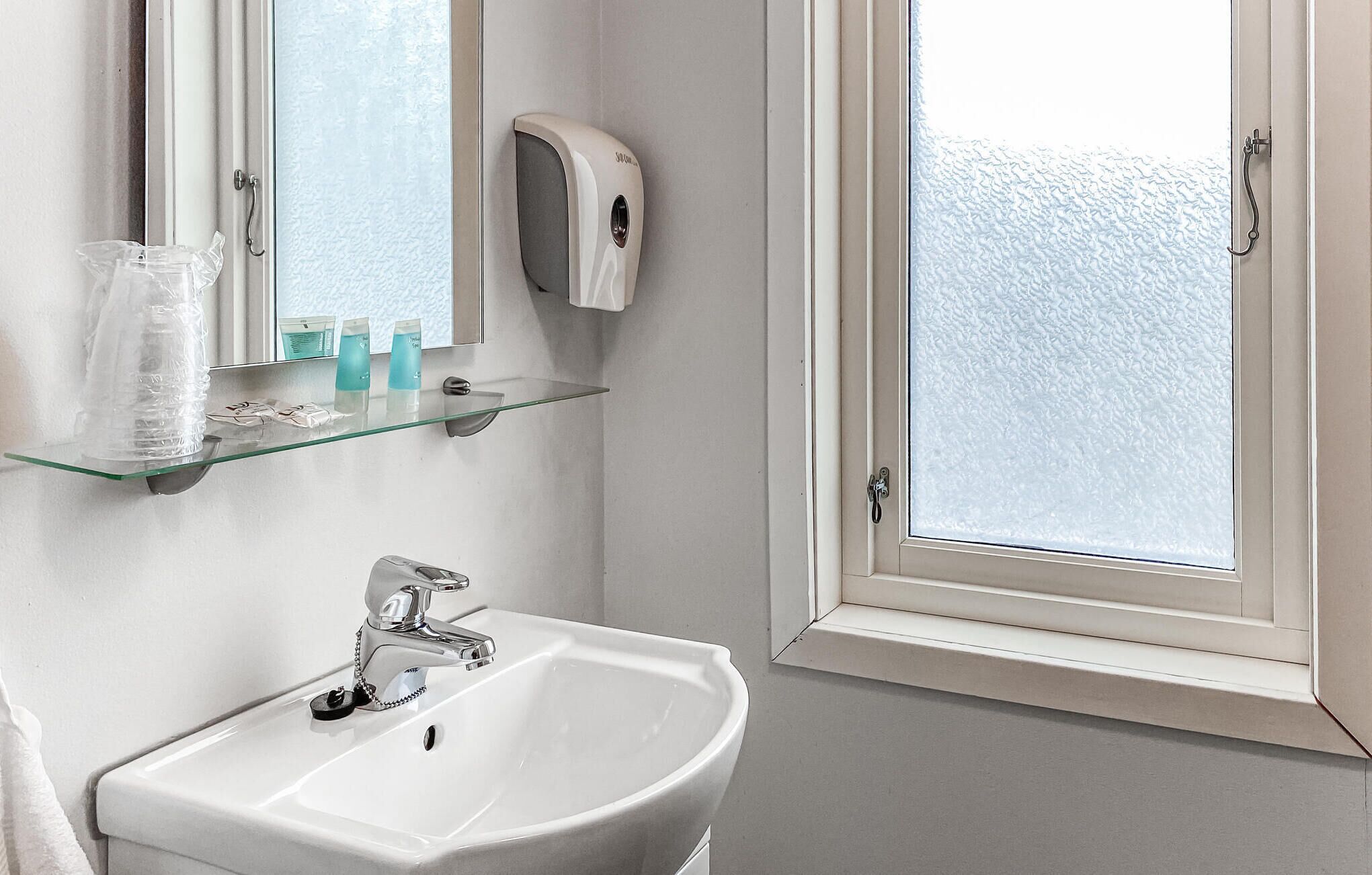 A bathroom with a white sink, a glass shelf with toiletries, a soap dispenser, and a frosted window.