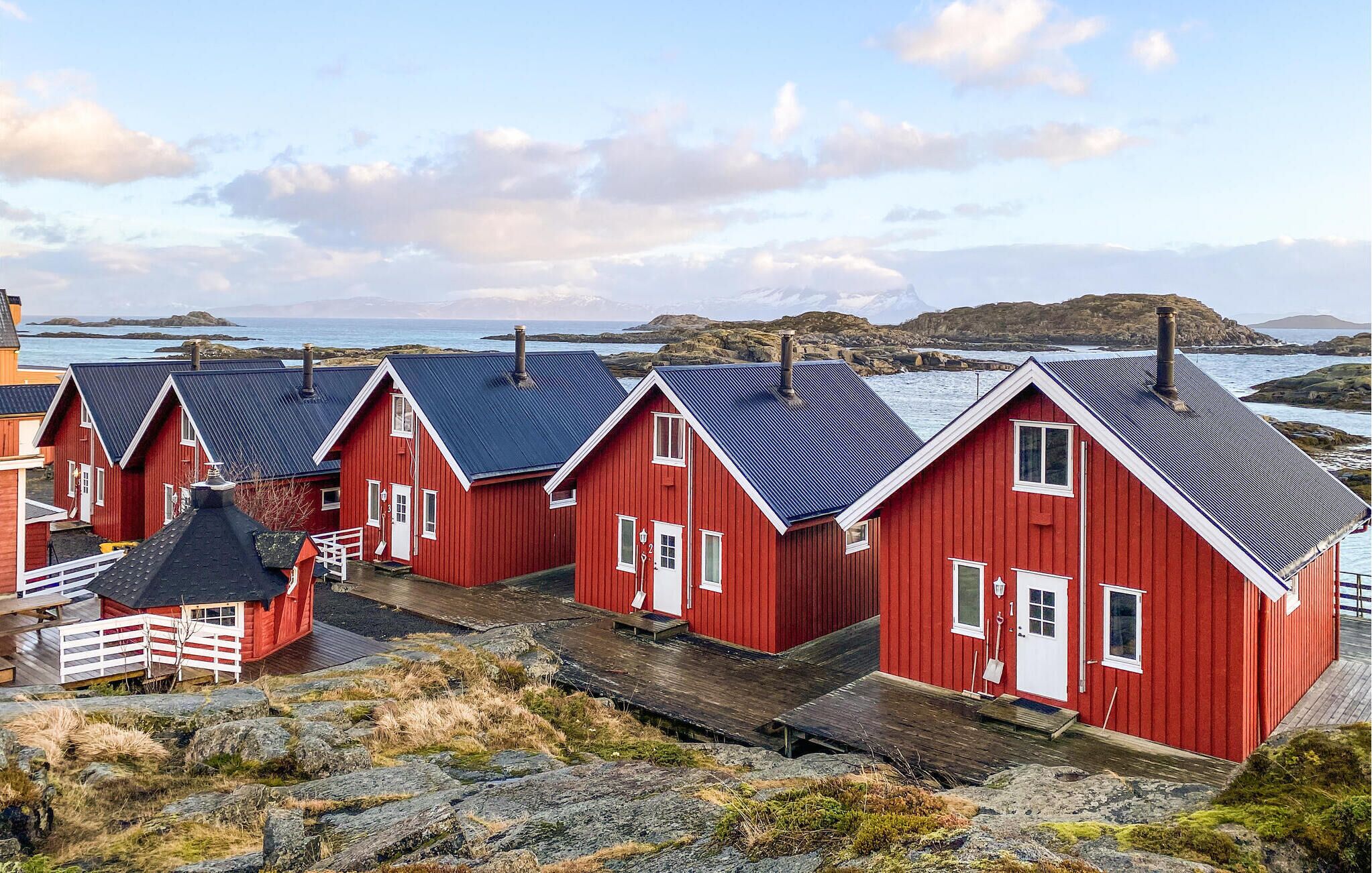 Una hilera de casas de madera roja con techos azules, detalles blancos y una costa rocosa con vistas al mar y a islas distantes.