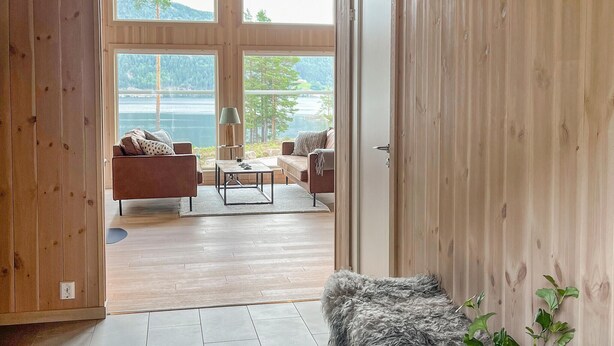 A room with wooden walls and floor, a sofa, a coffee table, and a lamp. There is a view of a lake and trees through large windows. - Lovely home in Treungen (Treungen)