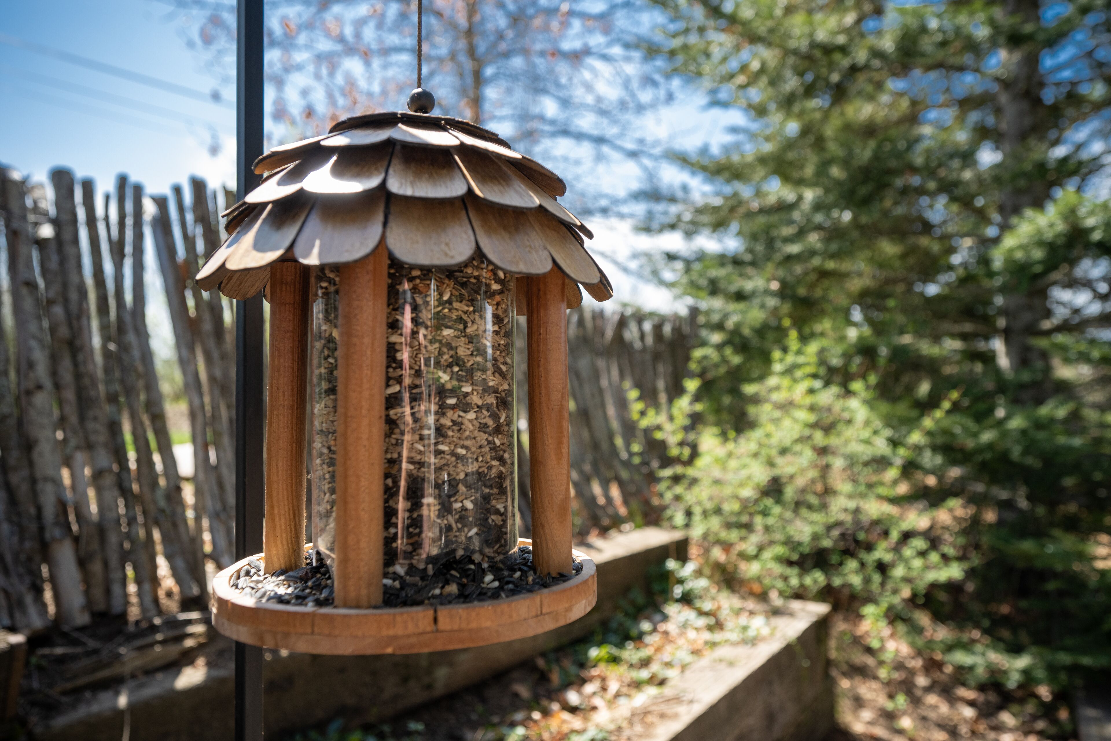 A bird feeder with a wooden structure and a circular base, surrounded by a wooden fence and greenery.