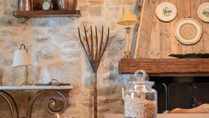 Un intérieur rustique avec un mur en pierre, une étagère en bois sur laquelle se trouve une horloge, une lampe en métal, un râteau en métal et un bocal en verre posé sur une table.
