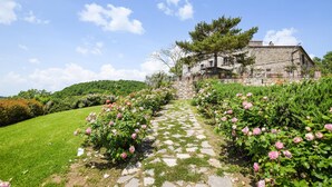 Une maison en pierre, accessible par un chemin de pierre, entourée d’une végétation luxuriante et d’arbustes en fleurs.