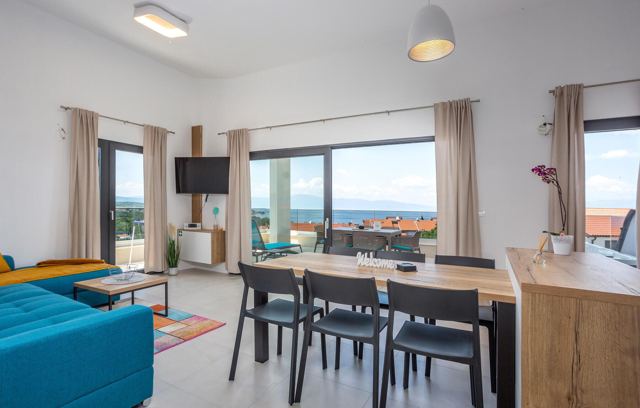 A modern living room with a sofa, dining table, chairs, a TV, and a view of the sea.