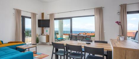 A modern living room with a sofa, dining table, chairs, a TV, and a view of the sea.