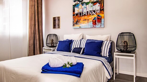 Una cama bien hecha con colcha a rayas blancas y azules, dos lámparas decorativas negras en mesitas de noche y un cuadro de barcos en la pared.