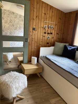 A cozy room with a wooden wall, a bed with a window, a small table, and a fluffy chair.