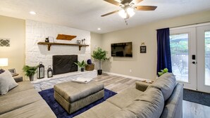 A living room with a stone fireplace, a ceiling fan, and a large window with curtains. - Backyard Family Oasis w/ Fire Pit + Deck, By Dwtn (Knoxville)