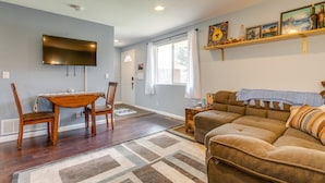 A living room with a brown sofa, a wooden dining table with chairs, a flat-screen TV mounted on the wall, and a window with curtains. - 12 Mi to Denver: Golden Vacation Rental! (Golden)