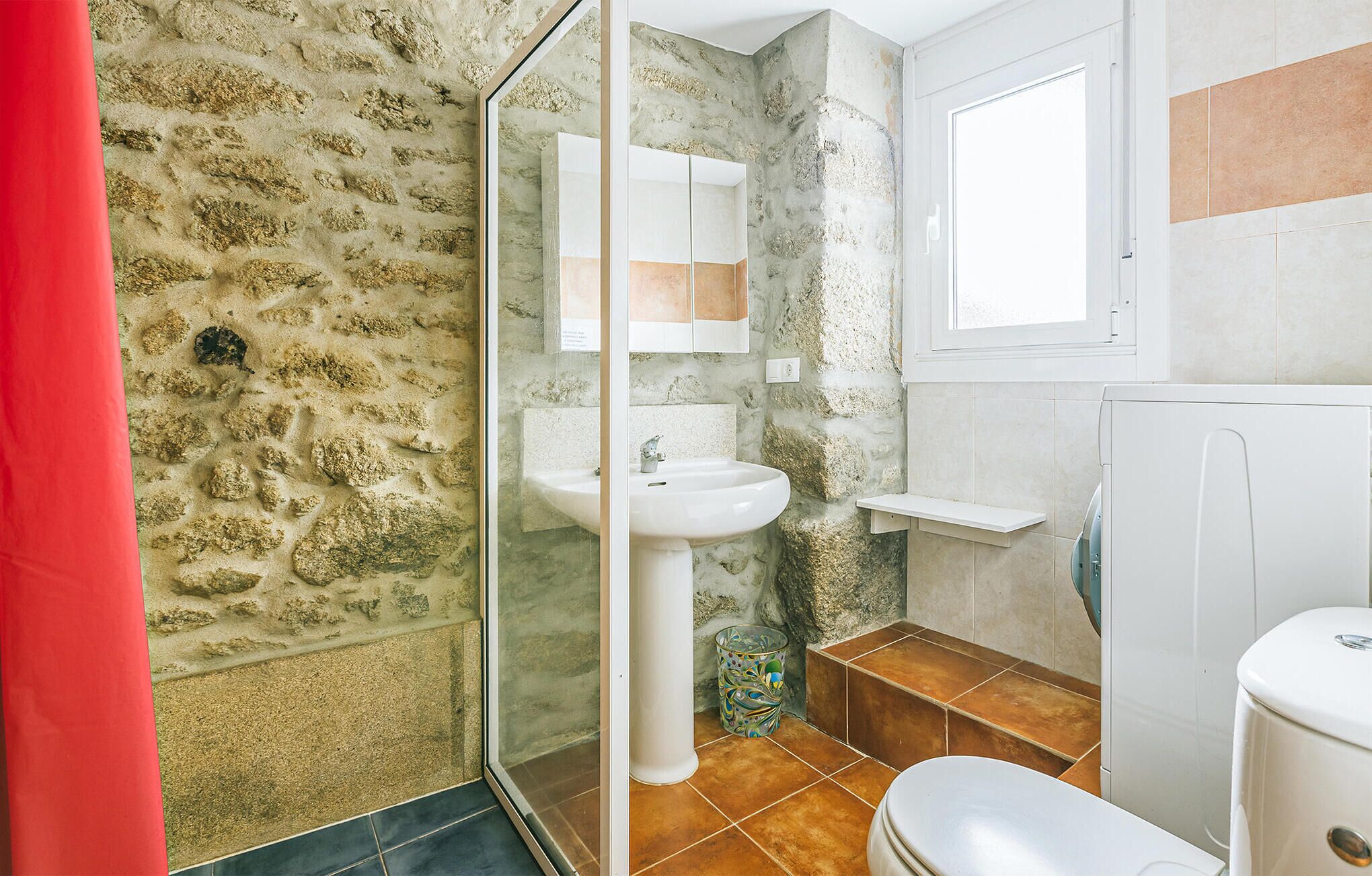 Een badkamer met een stenen muur, een glazen douchecabine, een witte wastafel en een toilet.