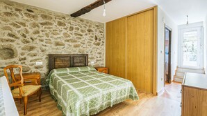 A bedroom with a stone wall, wooden bed frame, green bedspread, wooden wardrobe, and a window with a view.