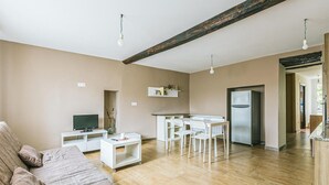 A living room with a brown sofa, a TV, a small white table, and a kitchen area with a fridge and a dining table.