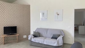 Olohuoneessa on harmaa sohva, jalustalla oleva televisio ja kaksi kehystettyä kuvaa seinällä.