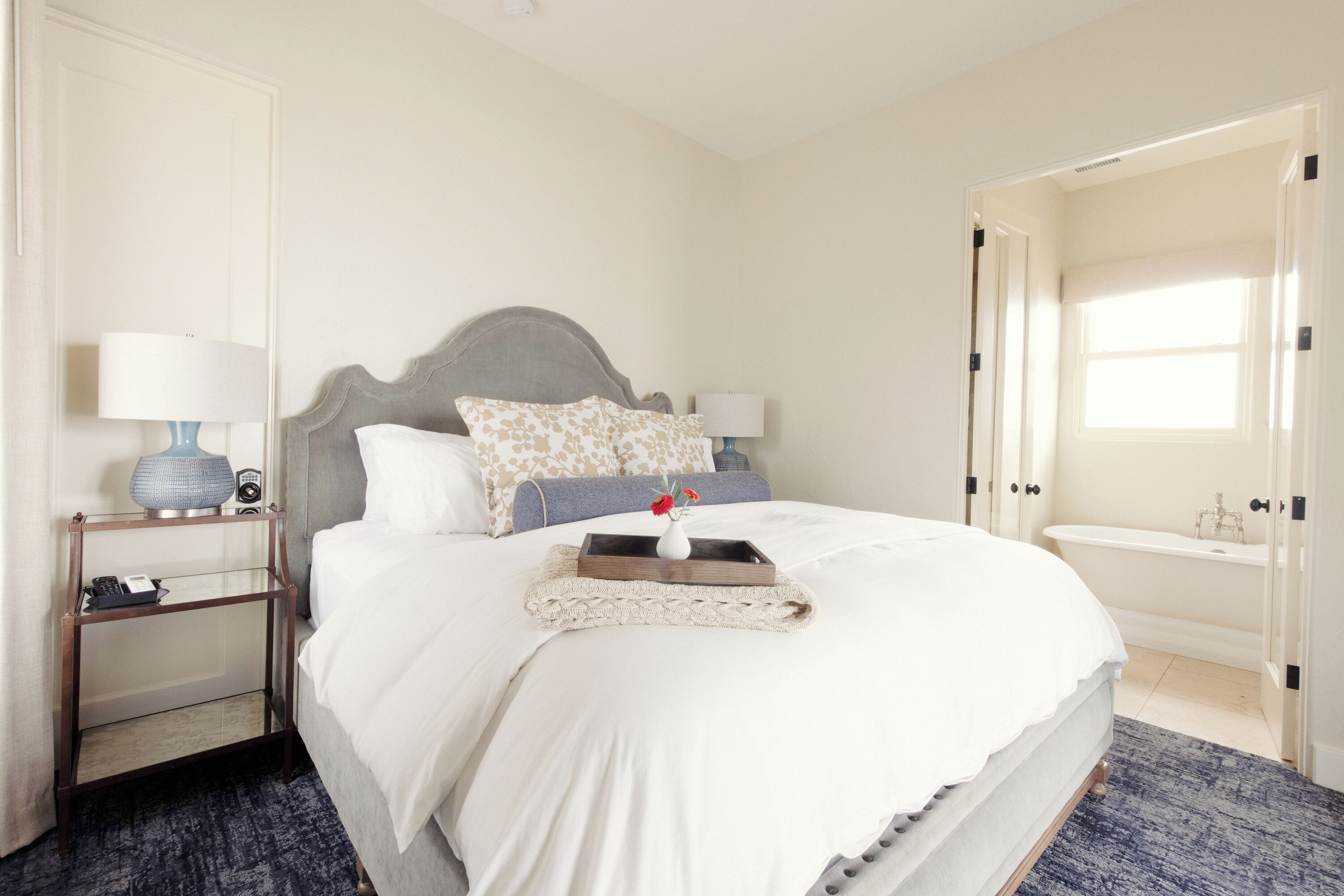 A neatly arranged bedroom with a bed, bedside tables, a nightstand, a tray with a vase, and a view of a bathroom.