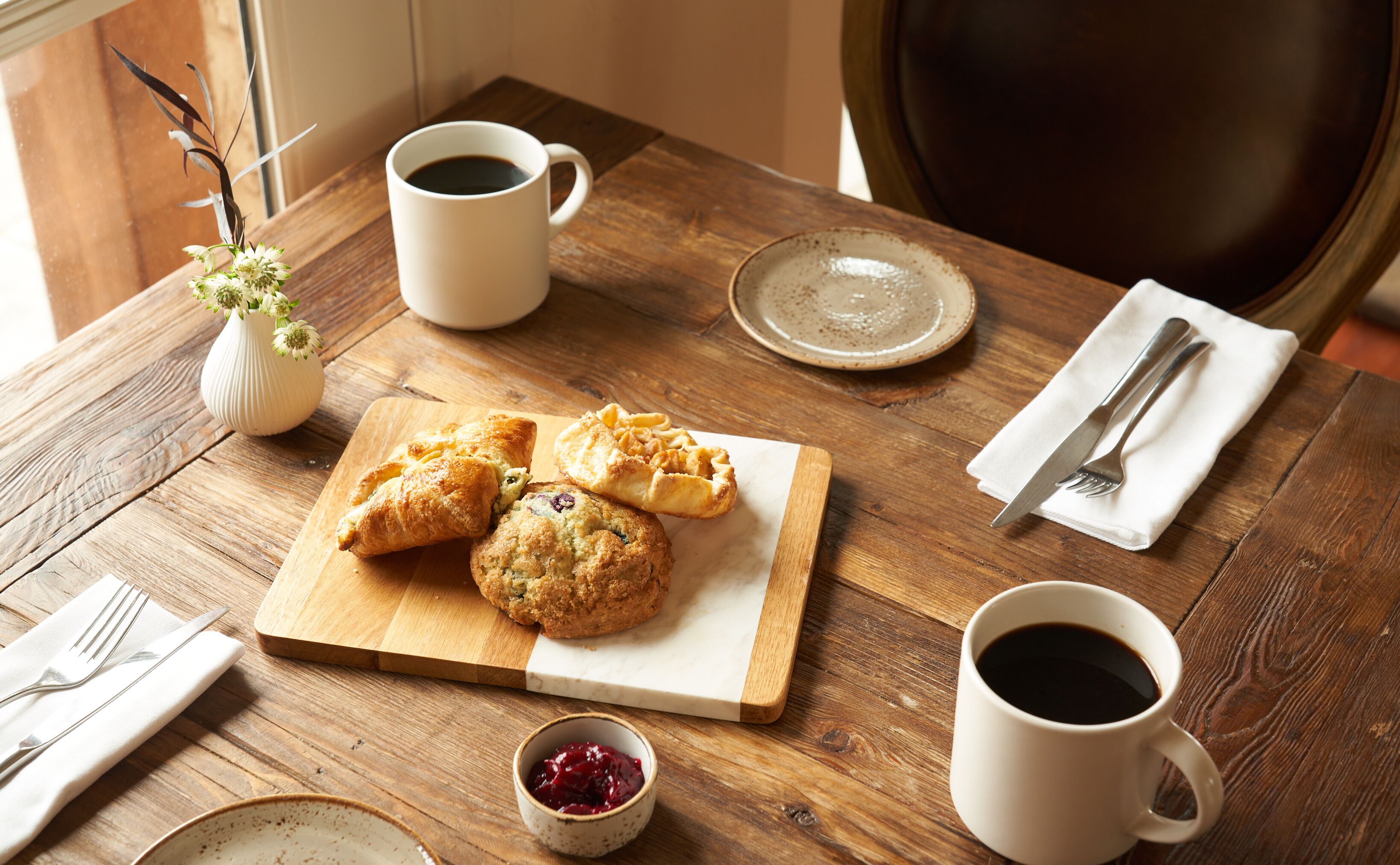 Una mesa de madera con una taza blanca, un plato de pasteles, un recipiente pequeño de mermelada y un florero con flores.