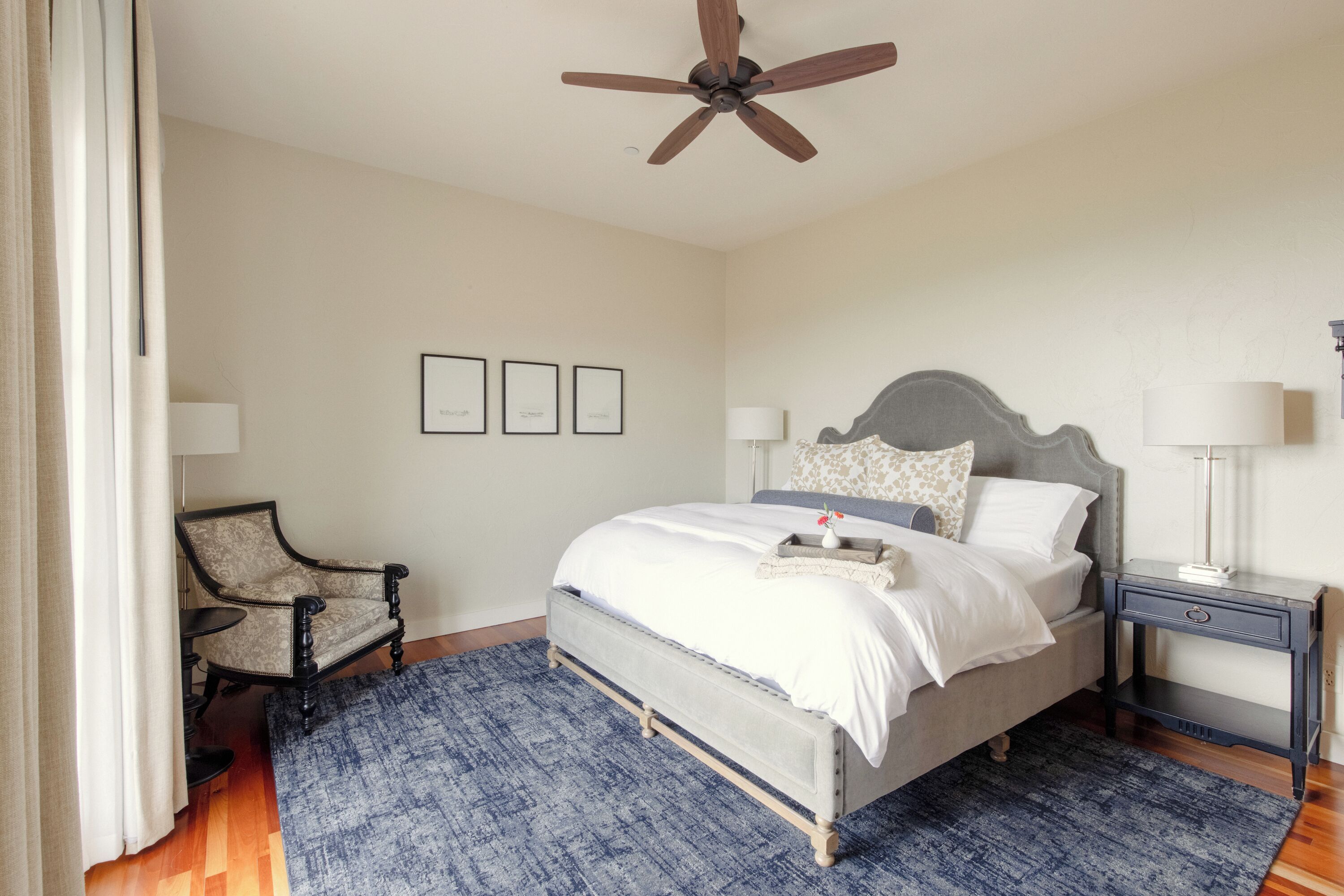 Ein Schlafzimmer mit Bett, Sessel, Nachttisch, Deckenventilator und gerahmten Bildern an der Wand.