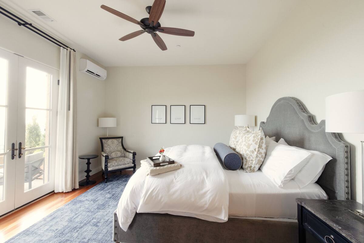 A bedroom with a bed, ceiling fan, air conditioning unit, framed pictures, and a chair.