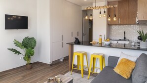 Een moderne keuken met een zwart aanrecht, houten kastjes en een ingebouwde oven. Er staan twee gele krukken en een grijze bank met een geel kussen. Een grote plant staat in de hoek, en een flatscreen televisie is aan de muur bevestigd.