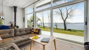 A modern living room with a large window overlooking a lake, a sofa, a coffee table with books, and a fireplace.