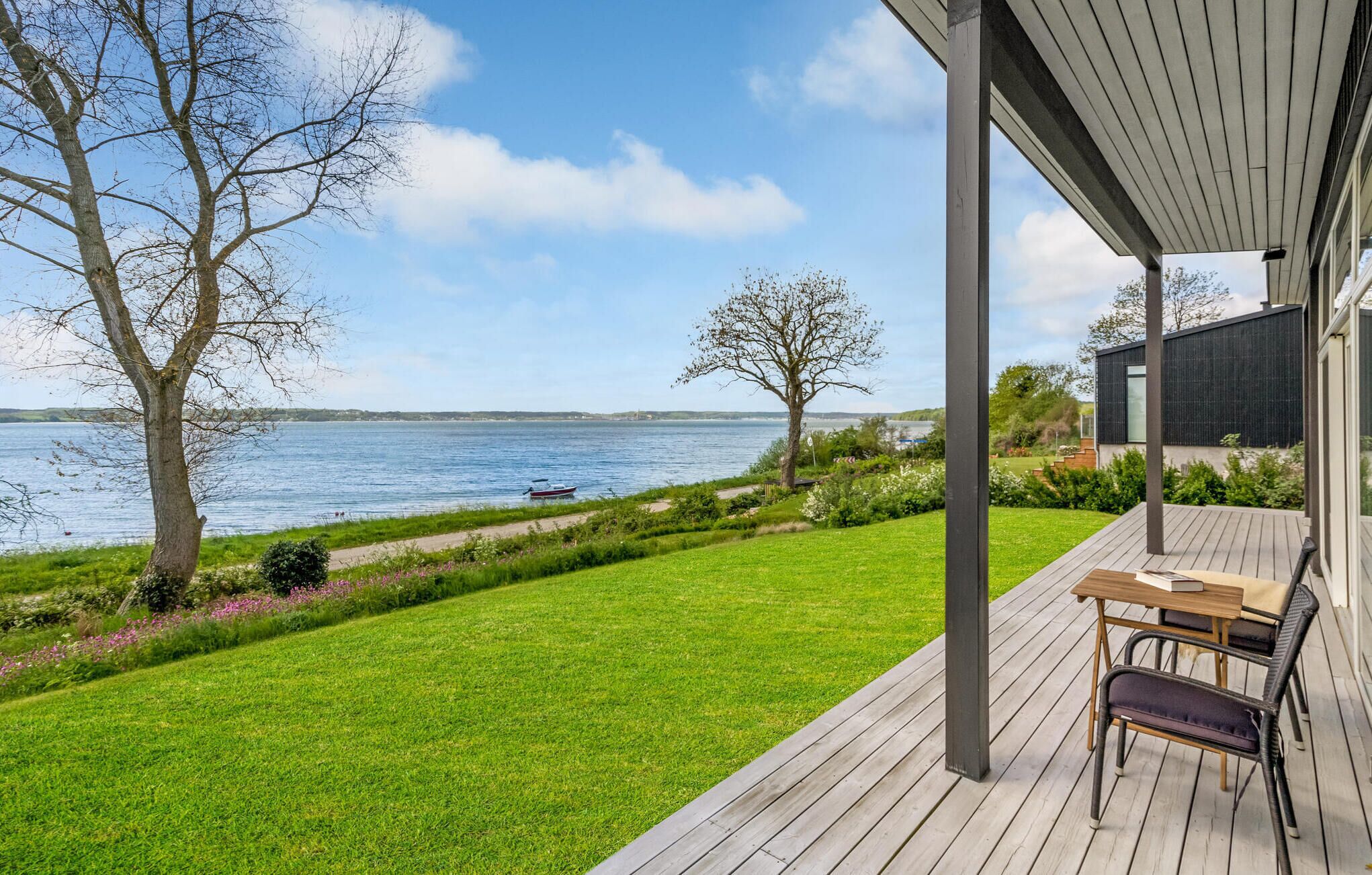 A wooden deck with a table and chairs, overlooking a lake with a boat and trees.