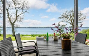 Une table avec des chaises, un vase rempli de fleurs, une bouteille verte et une carafe, le tout donnant sur un lac bordé d’arbres et sous un ciel dégagé.
