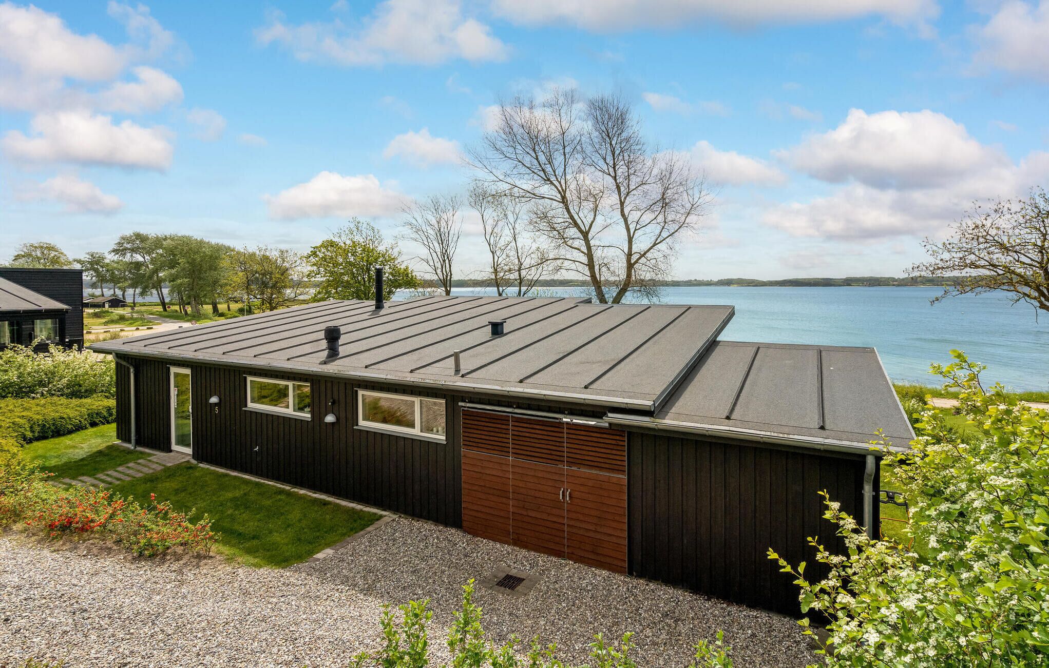 A modern building with a flat roof, large windows, and a garage door. It is situated near a body of water with trees and a clear sky in the background.