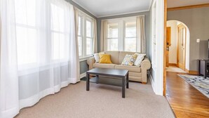A living room with a beige sofa, a dark coffee table, and sheer white curtains. - Home of the Braves - Bradley Campus Suite (West Peoria)