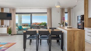Eine moderne Küche mit einer Holzinsel, einem Essbereich mit vier Stühlen, einem an der Wand montierten Fernseher und Meerblick durch große Fenster.