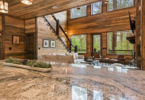 A spacious living area with a marble countertop, wooden walls, a staircase, and large windows offering a view of the forest.