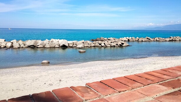 A beach with clear blue water, a rocky breakwater, and a brick pathway leading to the shore.