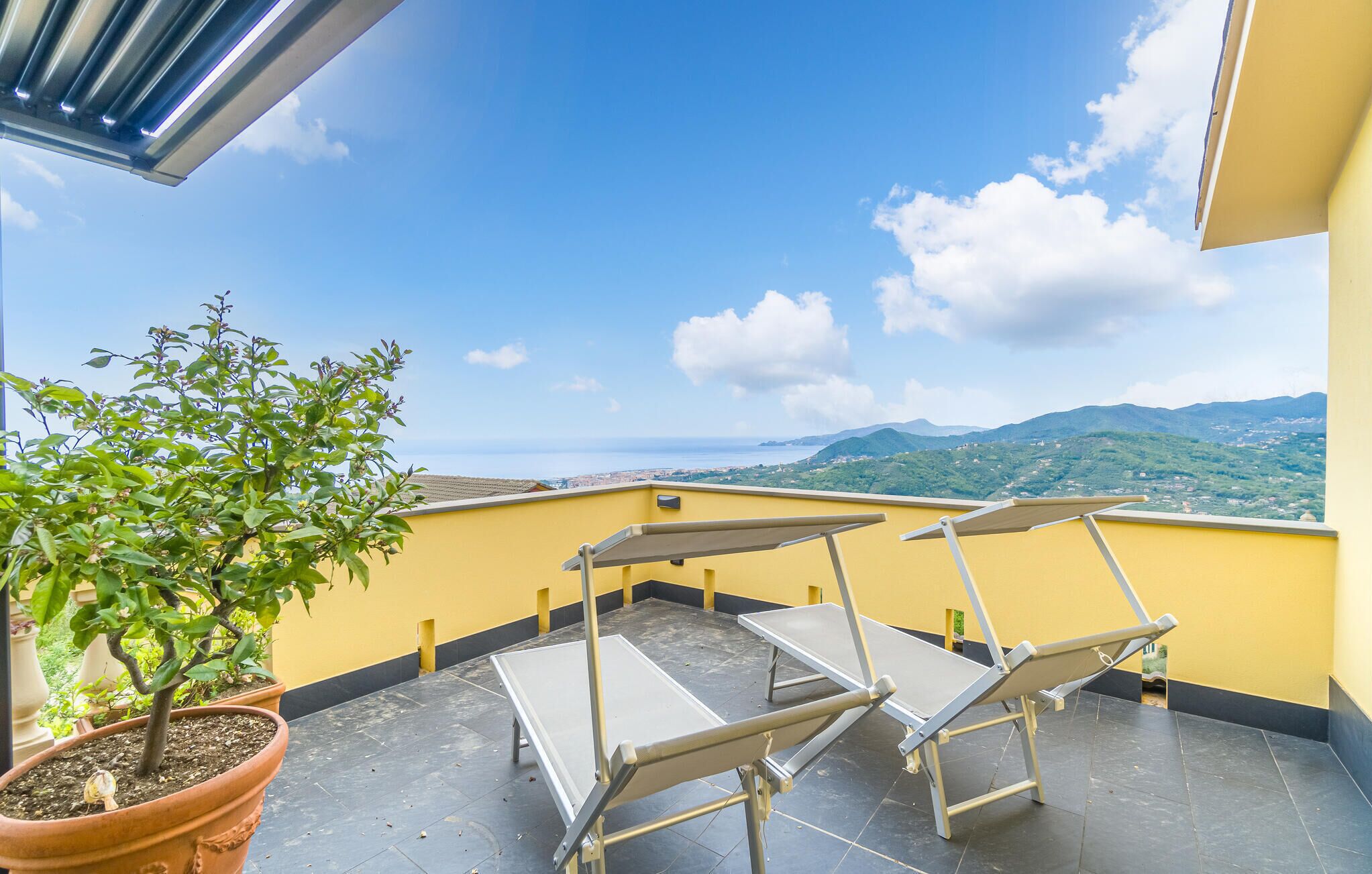 A balcony with two reclining chairs and a potted plant, overlooking a scenic view of mountains and the sea.