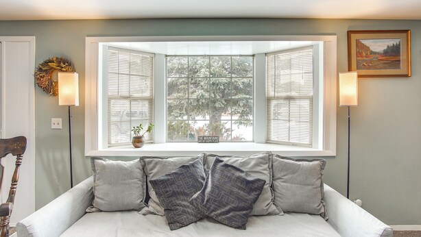 A light gray sofa with patterned pillows, a window with open shutters, and a framed painting on the wall.