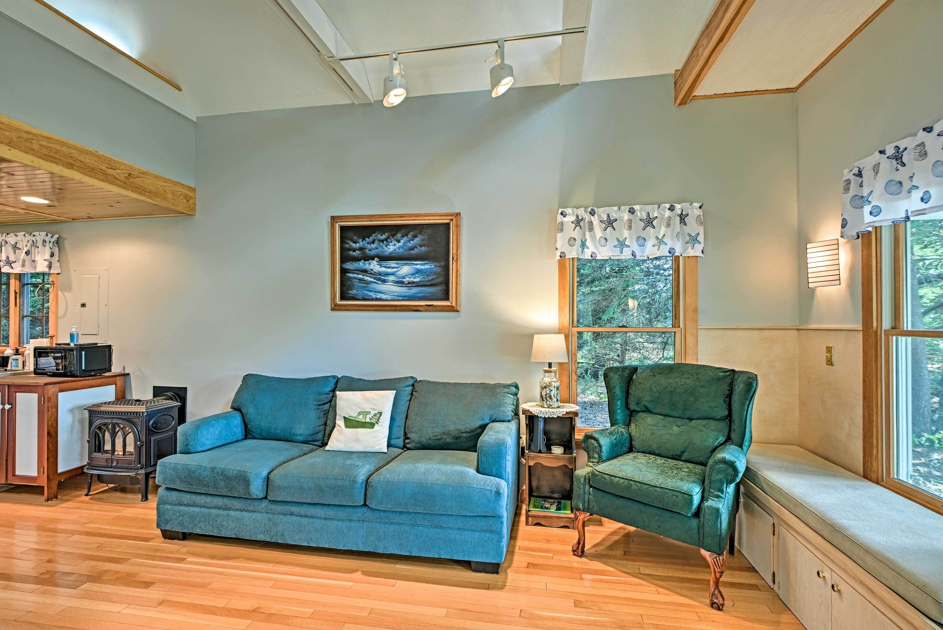 A cozy living room with a blue sofa, a green armchair, a wooden table with a microwave, and a painting on the wall.