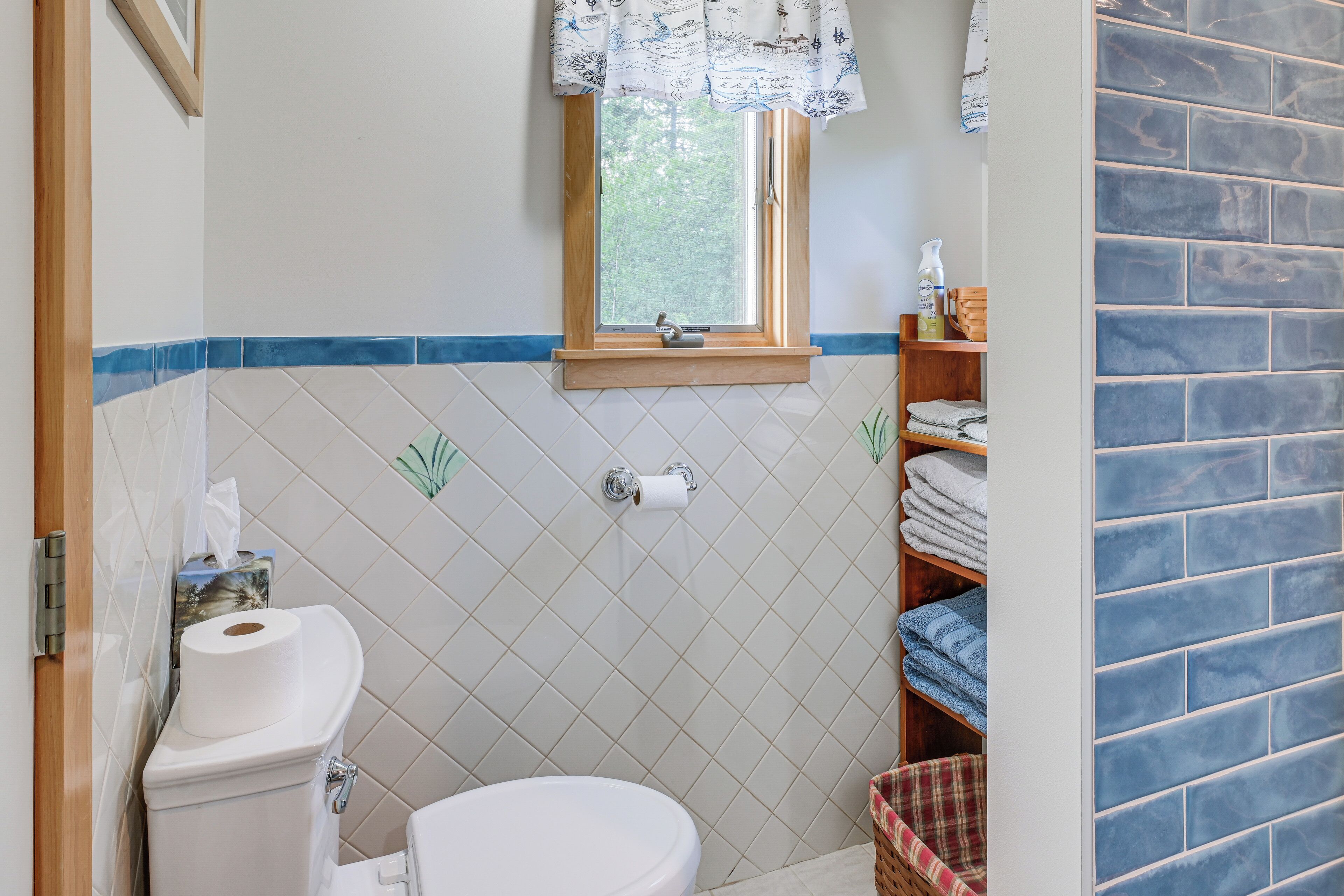 Une salle de bain avec des toilettes, une fenêtre avec un rideau, une étagère avec des serviettes et un mur carrelé.