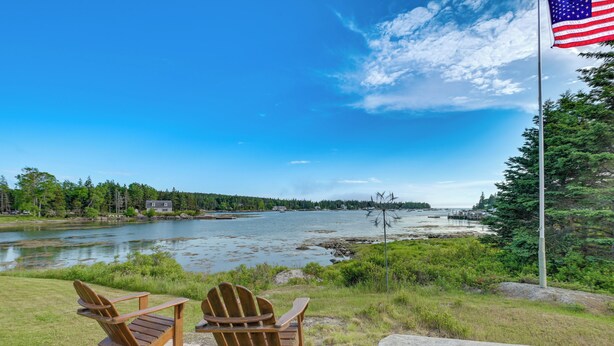 Deux chaises Adirondack en bois, placées face à une rivière, avec un mât de drapeau et un banc au premier plan.
