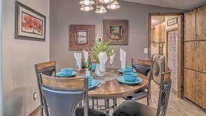 Un coin repas avec une table ronde, des chaises et un lustre. Des tableaux encadrés ornent le mur et une plante est posée sur la table.