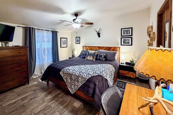 A bedroom with a bed, a ceiling fan, a dresser, a desk with a lamp, and a mounted deer head. - 'raven Hill Retreat' ~ Close to Midtown! (Ruidoso)