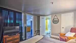 Ein Schlafzimmer mit Bett, Fernseher auf einer Kommode, einer Tür zu einem Badezimmer sowie Blick auf eine Küche und ein Wohnbereich.