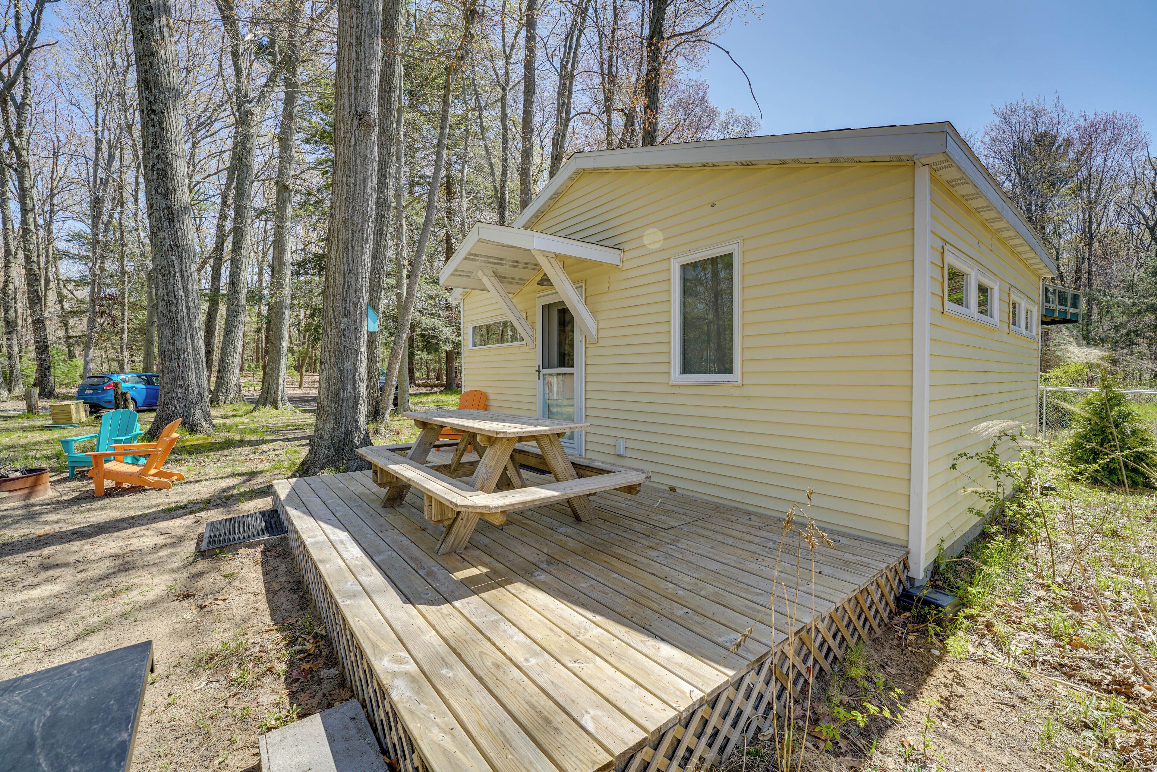 A small yellow cabin with a wooden deck, picnic table, and blue and orange chairs.
