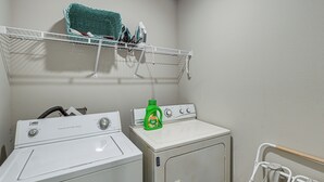 A laundry area with a front-loading washer and dryer, a shelf above with laundry items, and a bottle of detergent. - 'camden Ocean Springs' House Near Beaches! (Ocean Springs)