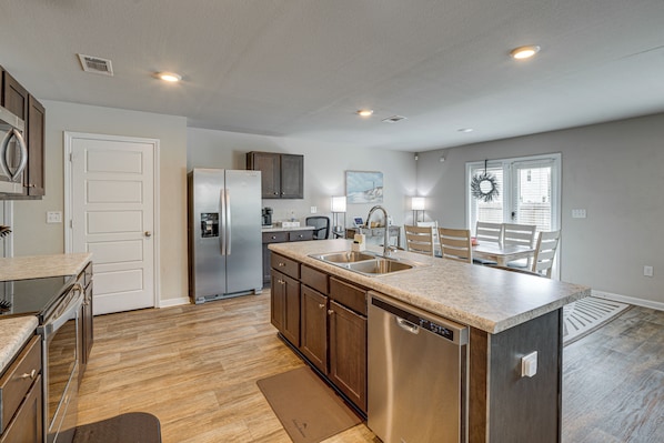 A modern kitchen with stainless steel appliances, a central island with a sink, wooden cabinets, and a dining area with a table and chairs.