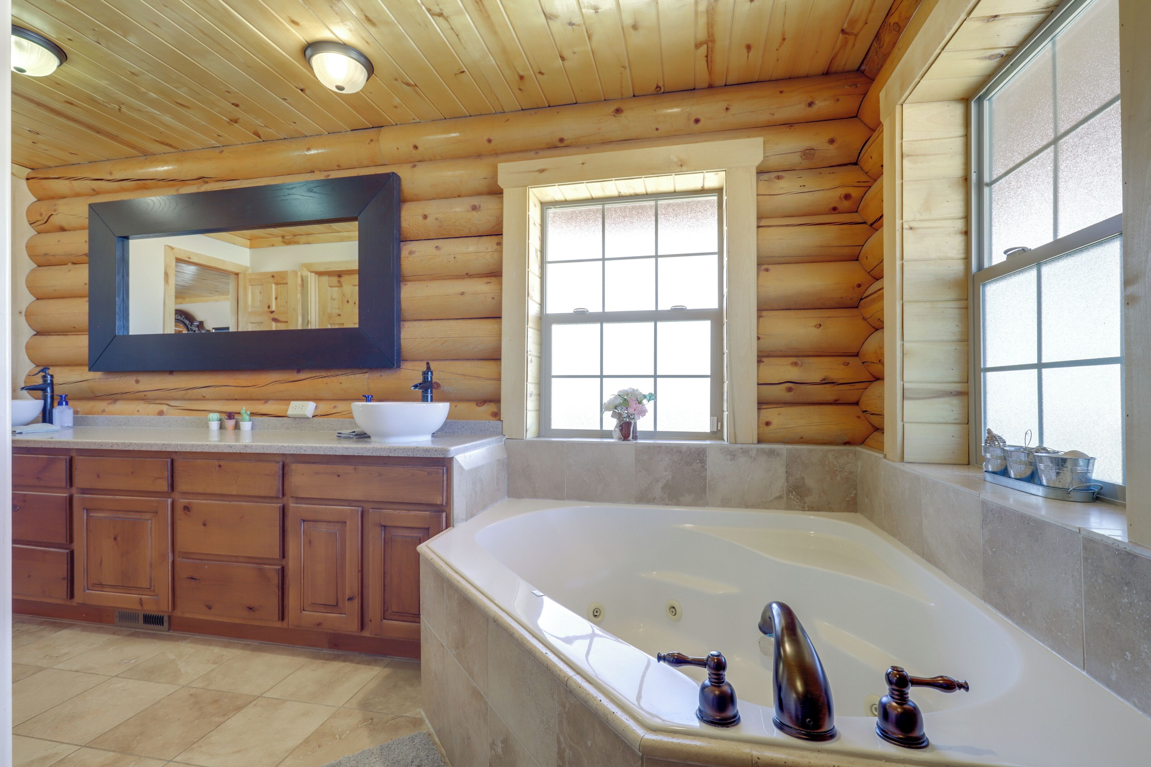 A log cabin bathroom with a large bathtub, wooden cabinets, a mirror, and a window.