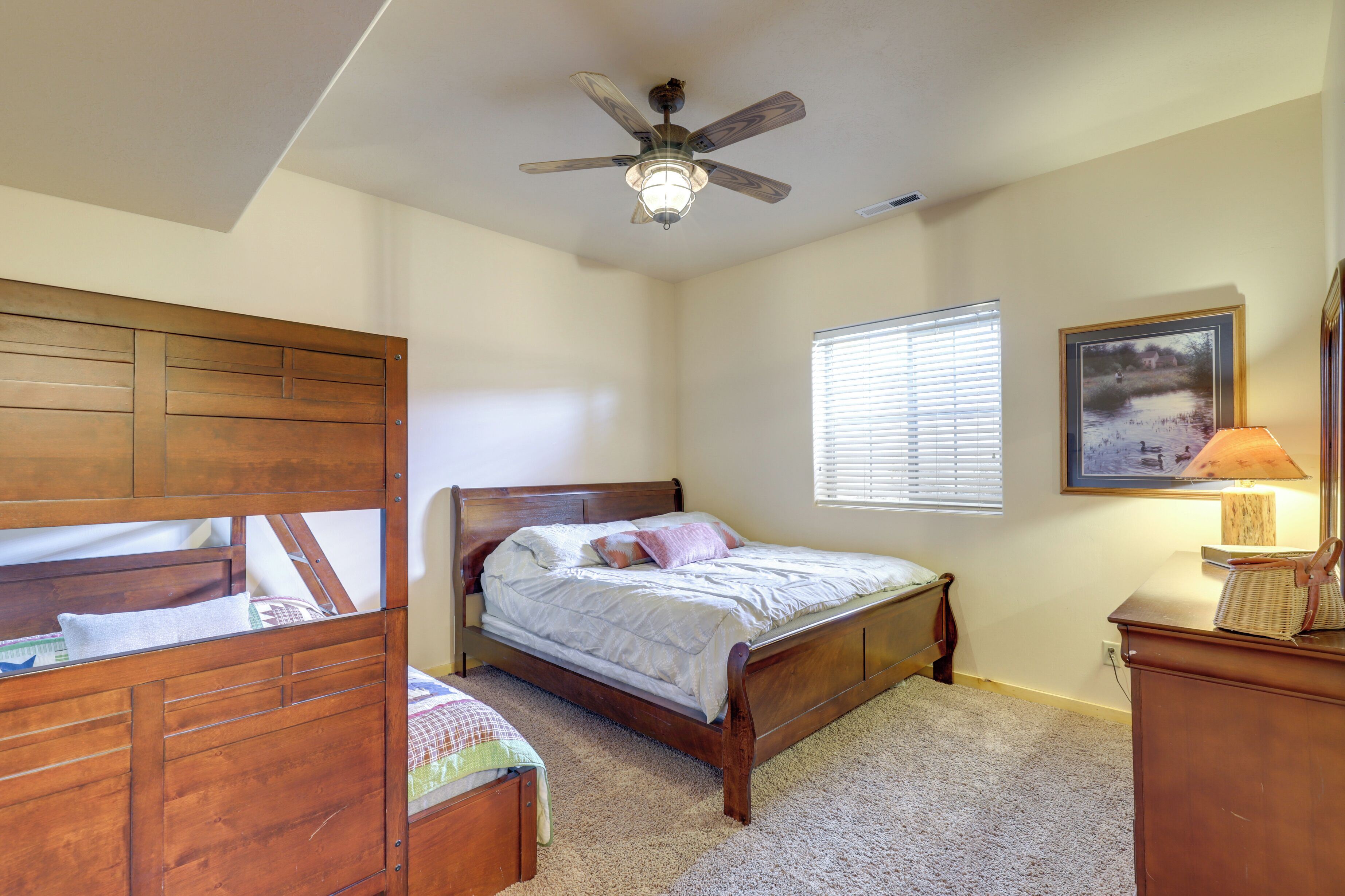 A bedroom with a wooden bed, a ceiling fan, a window with blinds, and a lamp on a dresser.