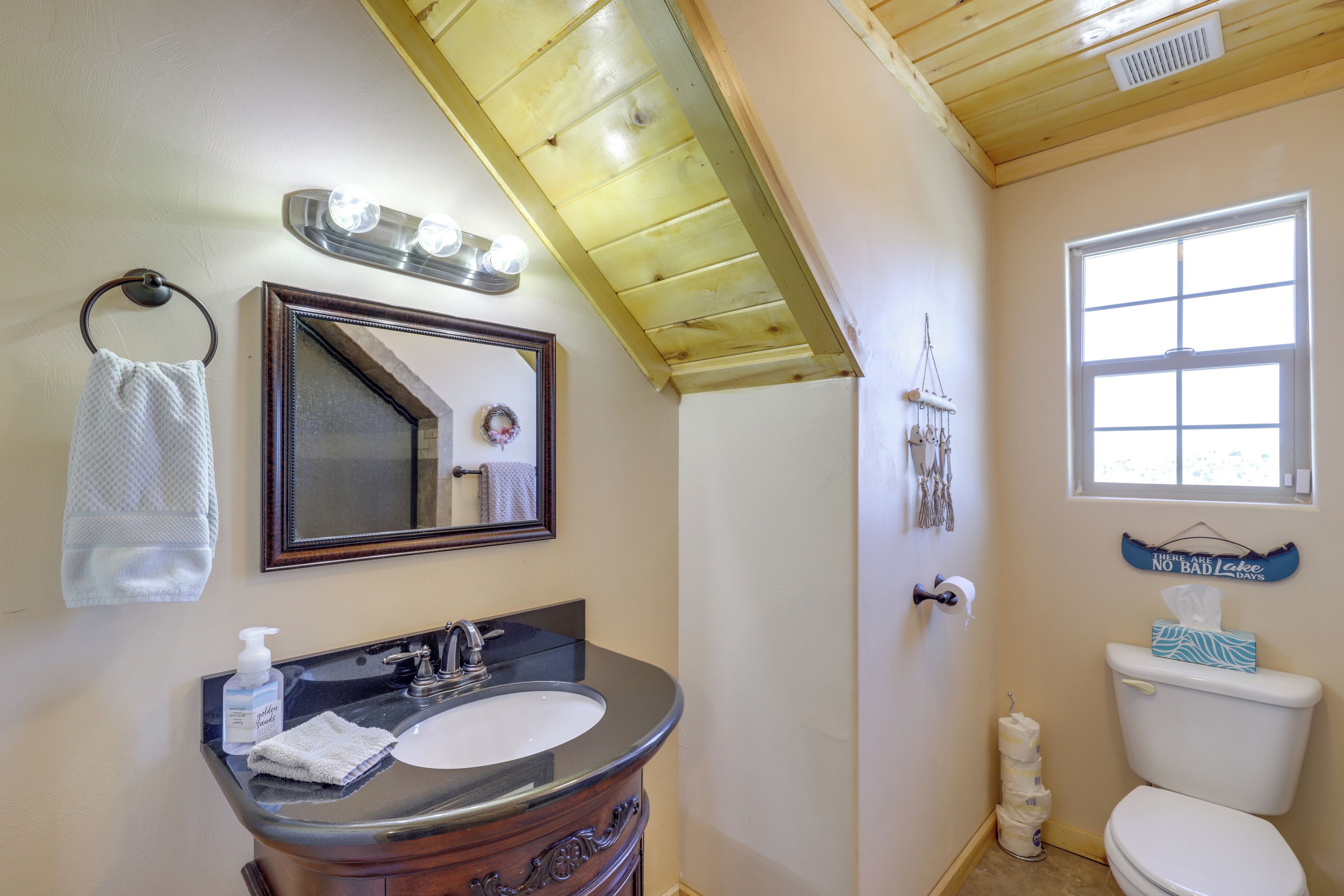 A bathroom with a wooden ceiling, a sink with a mirror, a toilet, and a window.