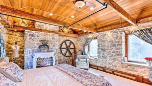 A rustic bedroom with a stone wall, a bed with floral bedding, a wooden ceiling, and a large window with curtains.