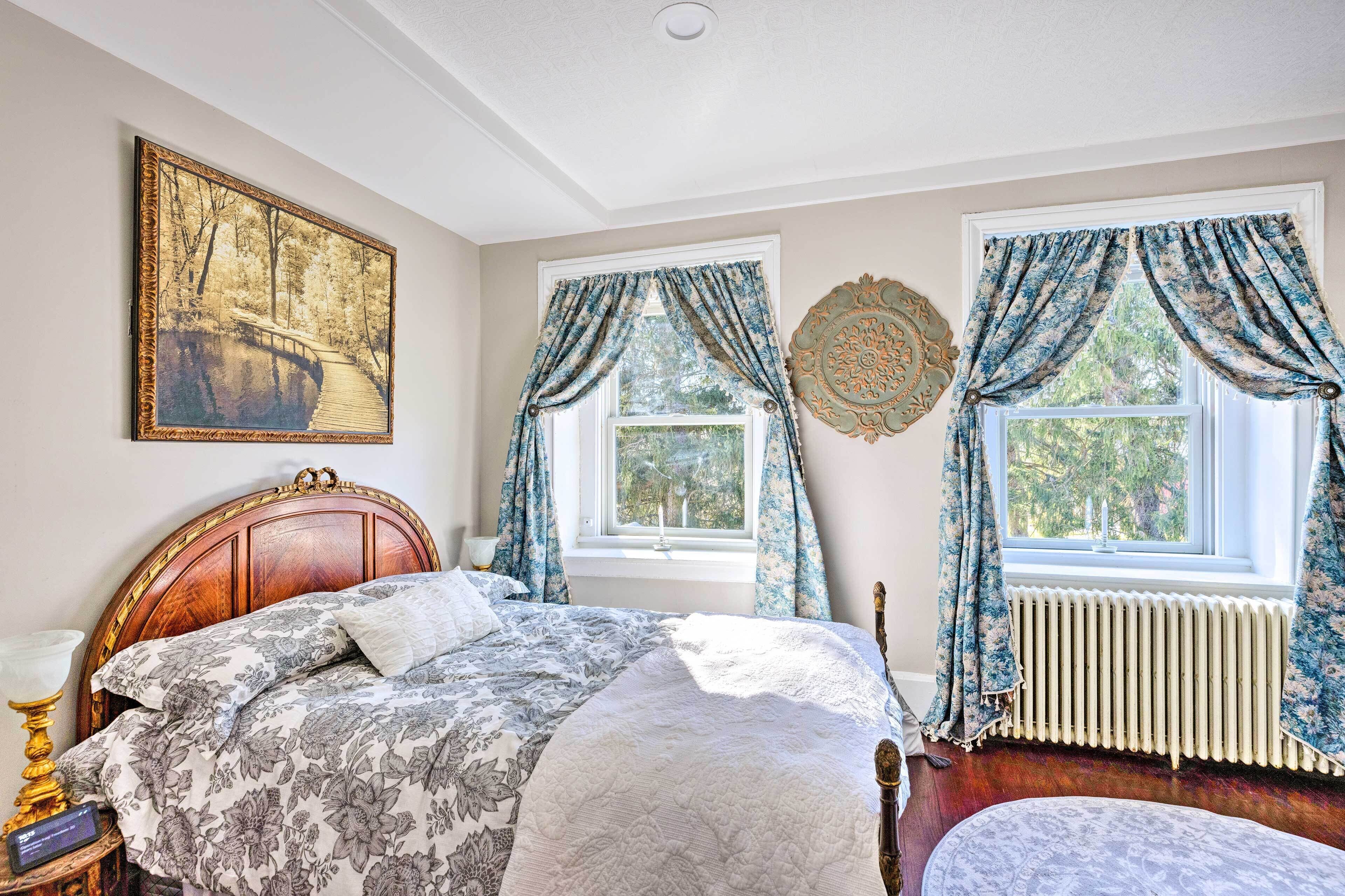 Een slaapkamer met een houten bed, een nachtkastje, een wandlamp, een raam met gordijnen, een radiator en een ingelijst schilderij aan de muur.