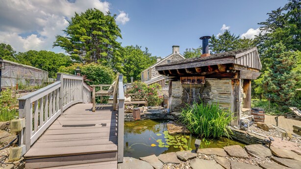 A wooden deck leading to a rustic stone building with a chimney, surrounded by lush greenery and a small pond with lily pads.